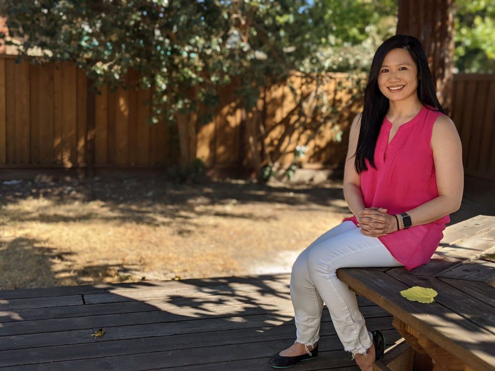 A photo of Alicia Chang sitting on a bench outside. She is looking into the camera and smiling.