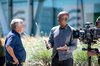 A behind-the-scenes view of Thomas Friedman and James Manyika filming their conversation, with glass windows and greenery from Google's Bay View campus in the background. Thomas, at left, is smiling and facing James, who is motioning with his hands and looking at Thomas. A person operating a camera is filming them in the foreground.