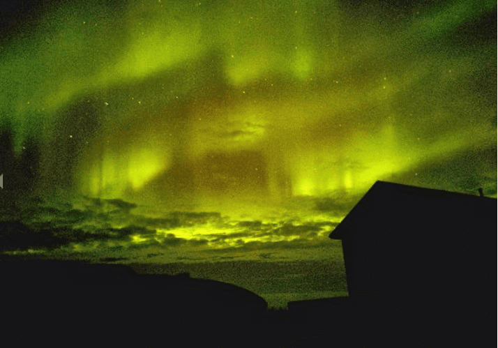 The aurora borealis, or northern lights, fill the night sky above a silhouette of a house..